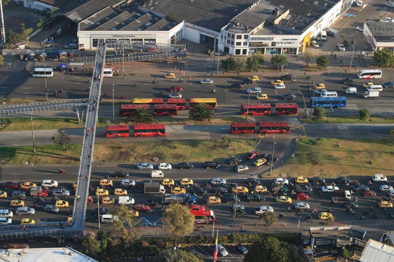 Una calle concurrida de la ciudad, se observan cinco carriles, varias transmilenios y carros masivos por las rutas