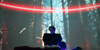 Imagen de un dj tocando en su tornamesa, de fondo se ve la imagen de un bosca iluminado con una luz roja de lado a lado.