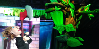 Imagen de un escenario en el que se encuentran un títere de un hombre vestido de chaqueta negra con una hacha en la mano frente a un gran árbol verde.