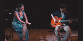 Imagen de una mujer sentada vestida de gala y de un hobre que toca guitarra en unescenario con fondo oscuro